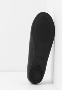 Semelle d'une chaussure noire avec grip texturé, taille 37, positionnée verticalement contre une surface blanche et un coin de mur.