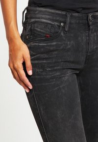 Schwarze Jeans aus Denim mit glatter Textur, schmaler Passform und subtilen Destroyed-Details. Enthält ein kleines rotes Logo auf der Gesäßtasche.