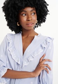 Blusa azzurro chiaro con dettagli ricamati, colletto e maniche arricciati. Tessuto morbido e vestibilità rilassata, con motivi floreali bianchi.