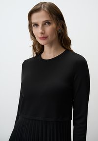 Haut noir à manches longues avec un buste ajusté et une jupe plissée. Texture lisse, col rond et détails de couture subtils aux épaules.