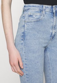 Jeans de mezclilla azul claro con una cintura alta, un bolsillo frontal con el logo de CK y un sutil desvanecimiento a lo largo de las piernas. Textura suave.