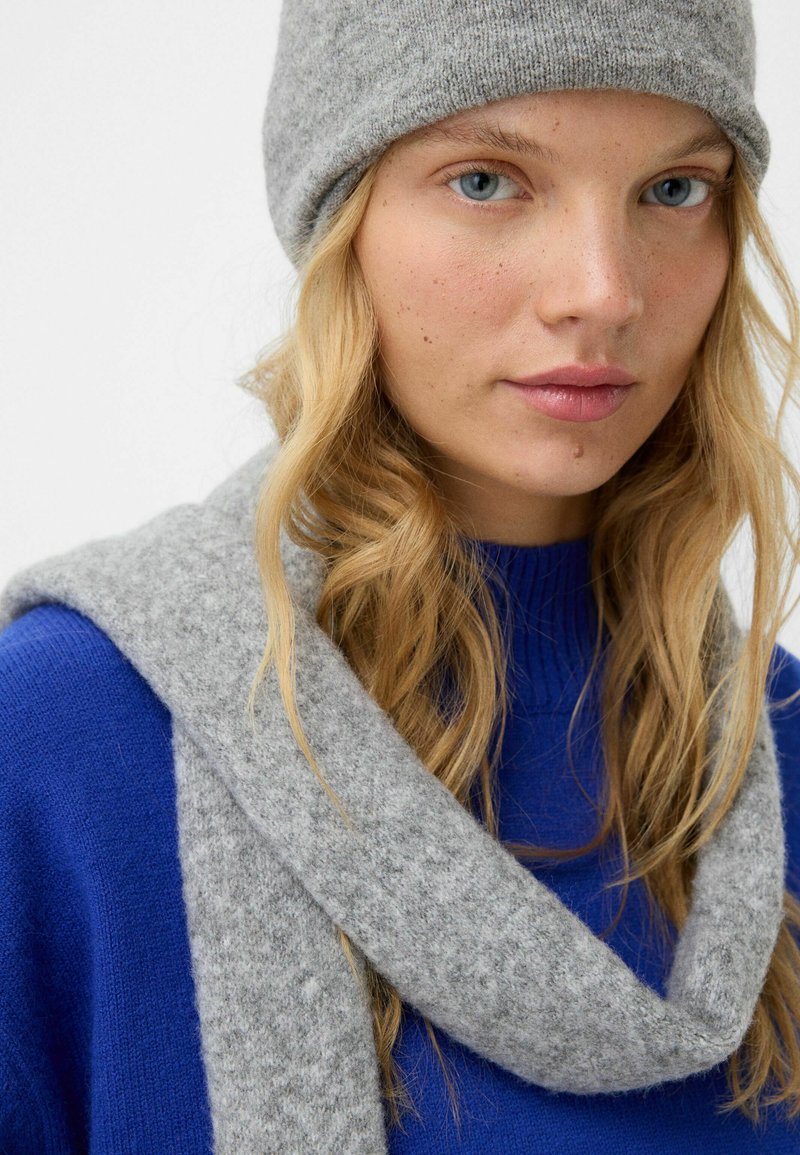 Un bonnet en tricot gris et une écharpe en tissu doux, portés sur un pull bleu. L'écharpe tombe autour du cou avec une texture lisse.