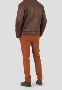 Uomo con una giacca di pelle marrone, pantaloni color ruggine e stivali scuri in suede, in piedi con le mani nelle tasche.