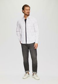 Witte shirt met een blauw en rood stippenpatroon, lange mouwen, knoopsluiting aan de voorkant, gecombineerd met donkere jeans en witte sneakers. Effen achtergrond.