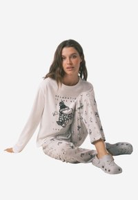 Witte pyjama top met lange mouwen en grafische print, gecombineerd met lichtgrijze broek met patroon met cartoonmotieven, en grijze instapschoenen.