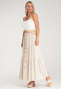 Beige Maxirock mit gestuften Design, Knopfdetails und einem geflochtenen hellbraunen Gürtel. Das Model trägt ein weißes geripptes Tanktop und neutrale Plateauschuhen.