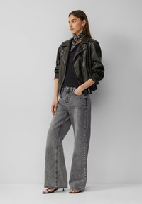 Zwarte leren motorjack over een zwart top, gecombineerd met grijze jeans met wijde pijpen. Zwarte sandalen en een gestreepte sjaal accentueren de look.