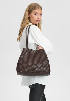 Blonde vrouw in witte shirt en zwarte broek draagt een grote donkerbruine geweven leren handtas over haar schouder terwijl ze omkijkt.