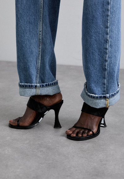 Sandale negre cu toc înalt, cu un design elegant, având o curea capitonată și o bandă subțire pentru degetul mare, asortate cu blugi din denim albastru cu manșete.