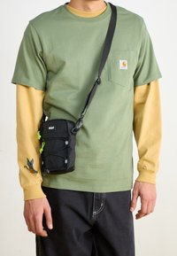 Persona que lleva una camiseta verde de Carhartt sobre una camisa de manga larga amarilla con un bordado de pato en la manga y un bolso cruzado negro.