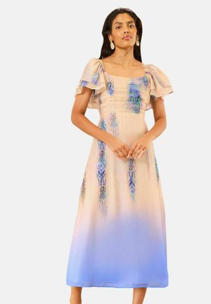 Kvinde iført en beige kjole med blå blomsterprint og puffede ærmer, stående med hænderne foldet foran mod en ensfarvet baggrund.