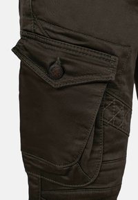 Poche cargo en tissu marron foncé sur un pantalon, avec rabat maintenu par un bouton rond et détails de double couture visibles.