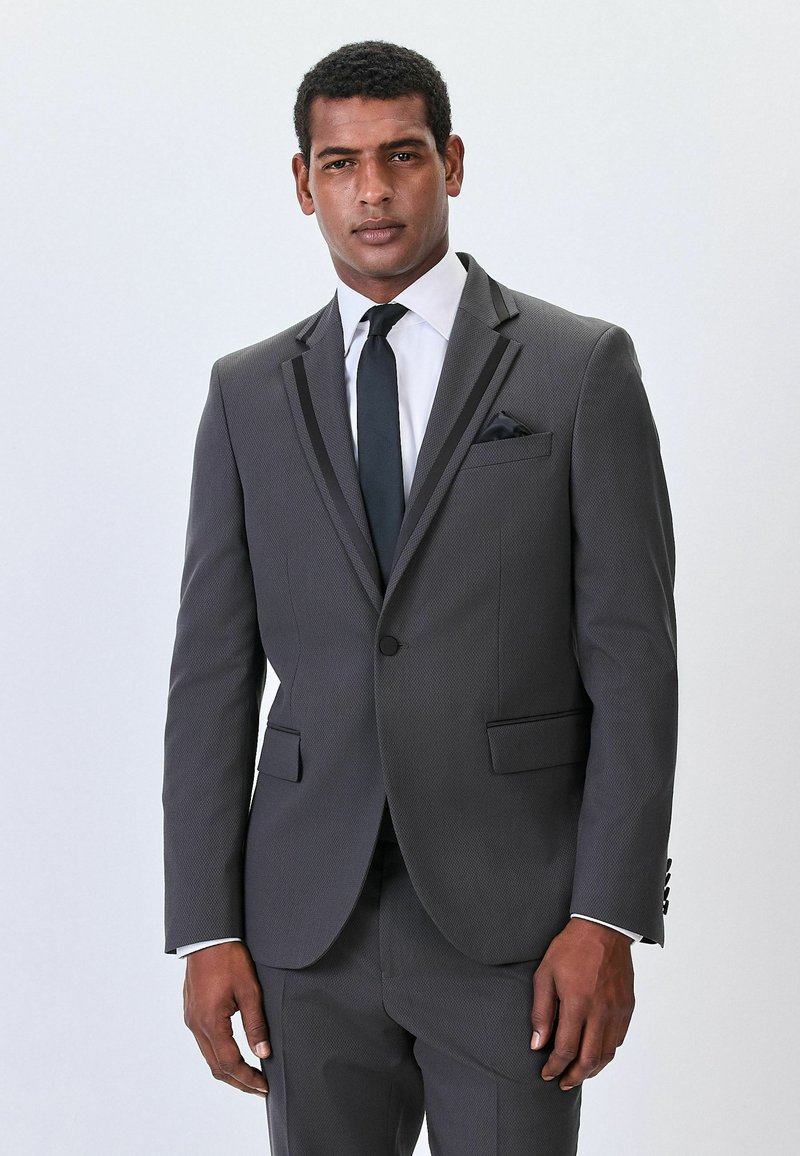 Traje gris con tejido texturizado, corte entallado, solapas en pico, ribetes de satén negro y corbata a juego. Incluye un pañuelo de bolsillo en el bolsillo del pecho.
