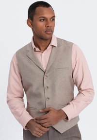 Chaleco texturizado beige con solapa de muesca, cinco botones y un bolsillo en el pecho, usado sobre una camisa de manga larga rosa clara.