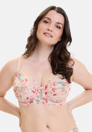 Mujer de cabello largo y oscuro que lleva un sujetador de encaje beige con patrones florales en rosa y verde, posando con las manos en la cadera sobre un fondo liso.