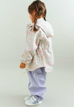 Ragazza giovane con capelli intrecciati che indossa una giacca con cappuccio floreale, pantaloni lucidi color lavanda e sneakers bianche, vista di profilo su uno sfondo neutro.