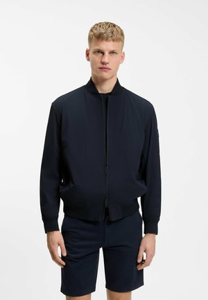 Ung mand med kort blondt hår iført en sort bomberjakke, der er lynet op, kombineret med sorte shorts, stående mod en hvid baggrund.