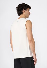 Giovane uomo con capelli ricci che indossa una maglietta bianca senza maniche e pantaloni corti neri, girato di lato su uno sfondo bianco semplice.