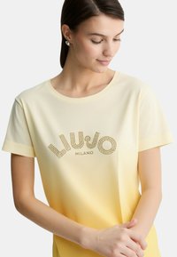 T-shirt giallo sfumato con maniche corte, caratterizzato da un logo "LIU JO MILANO" in strass, tessuto morbido e scollatura a girocollo.