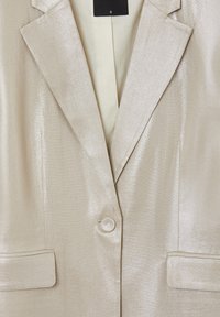 Blazer beige métallisé avec une finition texturée, revers crantés, fermeture à un bouton et deux poches avant. La doublure est de couleur crème.