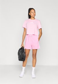 Rosa kort bomulls-T-skjorte med logo, matchende rosa shorts med snor og hvite detaljer. Modellen holder en svart treningsbag.