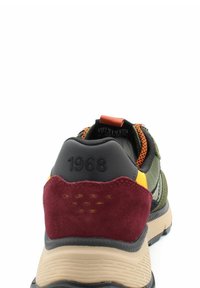 Sneaker da uomo con tomaia in tessuto verde oliva, tallone in suede bordeaux, accenti gialli e suola testurizzata. Presenta occhielli arancioni e un'etichetta per il trasporto.