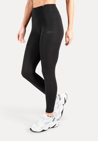 Zwarte leggings met hoge taille en een gladde textuur, voorzien van een subtiel logodetail op de dij, gecombineerd met witte sportsneakers.