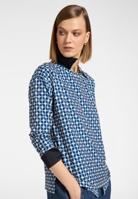 Blauw geruit overhemd met een ronde hals, lange mouwen en marineblauwe manchetten. Heeft een rasterpatroon in verschillende tinten blauw.