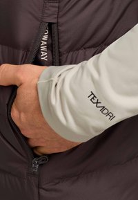 Donkergrijze geïsoleerde jas met een zichtbare rits en een hand die in een zak reikt. De mouw heeft een lichtgrijze stof met "TEXADRI" branding.