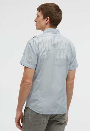 Junger Mann trägt ein hellblaues kurzärmeliges Hemd mit verblasstem Text auf dem Rücken und graue Hose, nach links gerichtet vor einfachem Hintergrund.