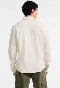 Mann mit kurzem, lockigem Haar, der ein cremefarbenes Langarmshirt und olivgrüne Cargo-Hosen trägt, mit dem Rücken zu einem weißen Hintergrund gewandt.