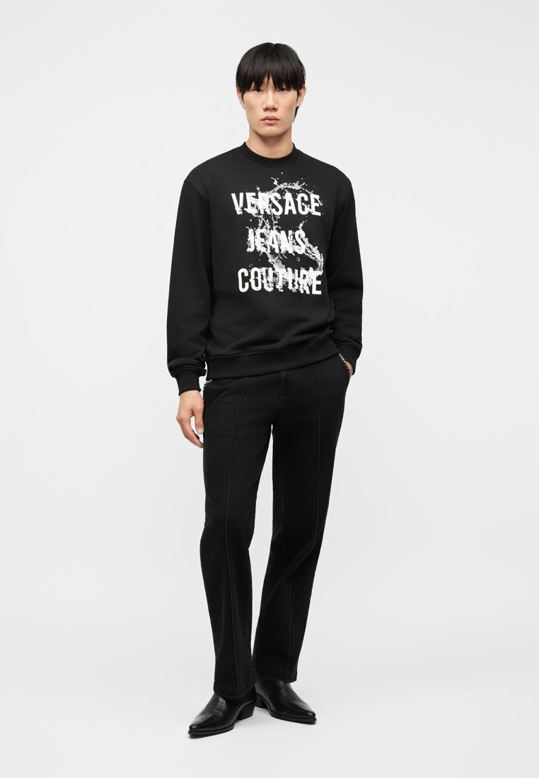 Jeune homme portant un sweat-shirt noir avec le texte blanc "Versace Jeans Couture" et un graphisme, un pantalon noir et des chaussures noires, debout devant un fond blanc.