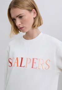 Vit sweatshirt med texten "SCALPERS" i gradientnyanser av rosa och rött, texturerad brodyr och rund halsringning.
