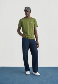 Πράσινο-ελιά βαμβακερό t-shirt με λογότυπο στο στήθος, συνδυασμένο με σκούρο μπλε τζιν και λευκά αθλητικά παπούτσια· τονίζεται με γκρι καπέλο.