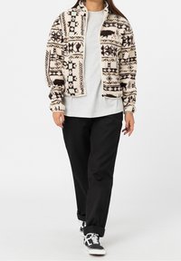 Futrzana kurtka w kremowym kolorze z geometrycznymi wzorami w czerni i niebieskim, z zapięciem na zamek i wysokim kołnierzem, noszona na szarej koszulce i czarnych spodniach.