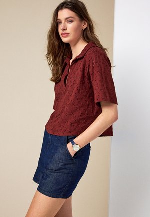 Junge Frau mit langen braunen Haaren, die eine burgunderrote Lochbluse und dunkle Denim-Shorts trägt und mit einer Hand in der Tasche steht.