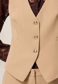 Chaleco beige ajustado con cuatro botones dorados, diseño estructurado y textura sutil. Debajo lleva una blusa de encaje oscuro de mangas largas.