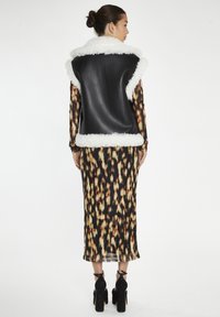 Gilet in ecopelle nera con rifiniture in pelliccia bianca, indossato su un vestito bodycon stampato con un motivo di tonalità nere, marroni e crema. Scarpe con tacco alto.