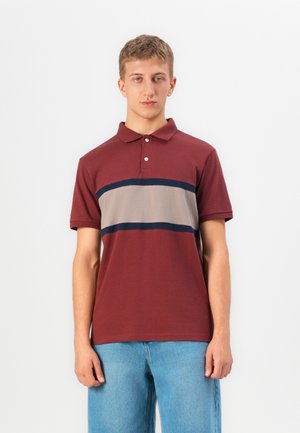 Ung man som bär en vinröd polo skjorta med beige och marinblå horisontella ränder samt ljusblå jeans, står framför en enfärgad vit bakgrund.