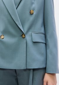 Ανοιχτό μπλε ραμμένο σακάκι με λεία υφή, με δύο μεγάλα κουμπιά από χελώνα και ένα θυλάκι τσέπης στην αριστερή πλευρά.