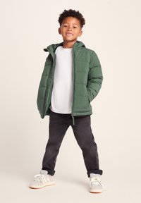 Green puffer jacket with a hood, quilted design, zip closure, worn over a white shirt, paired with dark jeans and gray sneakers.