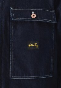 Riflová bunda z modré látky, kontrastní bílé švy, náprsní kapsa s měděným nýtem a černá visačka s textem ve zlaté barvě.