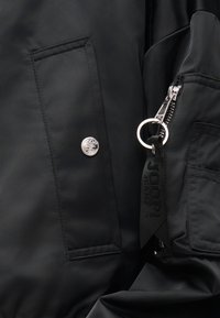 Ύφασμα μαύρου μπουφάν που δείχνει ένα κουμπί τύπου snap με λογότυπο και μια ταινία φερμουάρ προσαρτημένη σε μια υφασμάτινη θηλιά με ανάγλυφο κείμενο.