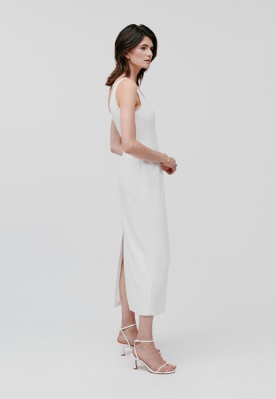 Witte, mouwloze, enkel-lange jurk met een zijsplit. Glad, aansluitend materiaal. Gepaarde met strapless witte haksandalen.