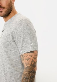 T-shirt henley à manches courtes rayé gris avec boutons, comprenant une poche et un détail de marque sur la manche. Texture douce et coupe décontractée.