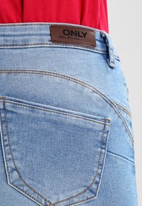 Jeans de mezclilla azul claro con cintura de media altura, que presentan una etiqueta de marca de cuero marrón y un bolsillo trasero con costuras en contraste.