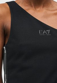 Μαύρο τοπ με έναν ώμο φορεμένο σε πρόσωπο με το λογότυπο EA7 Emporio Armani στο στήθος και λευκή διακοσμητική ταινία με το εμπορικό σήμα κατά μήκος της πλαϊνής ραφής.