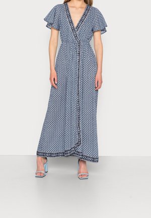 Vrouw die een blauwe wikkelmaxi-jurk met patroon draagt, met afhangende mouwen en lichtblauwe open-toe blokhakken, staand tegen een effen achtergrond.