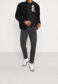 Chaqueta de pana negra con cremallera frontal, combinada con una camiseta gráfica negra y unos vaqueros pitillo gris oscuro. Zapatillas blancas con rayas negras.