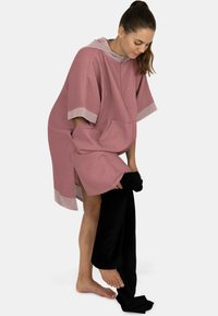 Rosa Kapuzen-Poncho aus strukturiertem Stoff, mit kurzen Ärmeln und einer Fronttasche. Das Modell zieht gerade schwarze Hosen aus, während es barfuß ist.